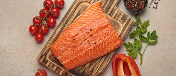 Taze somon fileto, ahşap kesim tahtasının üzerinde biber, kiraz domatesleri, kırmızı dolmalık biber ve maydanozla birlikte. Bej arka planda sağlıklı yemek malzemeleri. Üst görünüm bileşimi