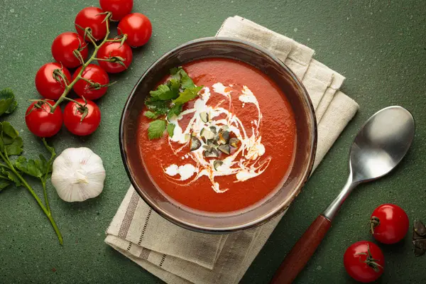 Taze domates çorbası krema, tohum ve otlarla dolu bir kasede servis edilir, yanında da yeşil bir zemin üzerinde yeşil bir kaşık ve peçeteyle kiraz domates, sarımsak ve maydanoz bulunur.