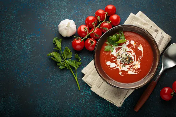 Taze domates çorbası krema, tohum ve otlarla dolu bir kasede servis edilir, yanında da mavi bir zemin üzerinde domates, sarımsak ve maydanoz, kaşık ve peçeteyle birlikte fotokopi alanı bulunur.