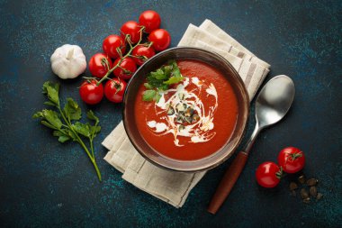 Taze domates çorbası krema, tohum ve otlarla dolu bir kasede servis edilir, yanında da mavi bir zemin üzerinde kaşık ve peçeteyle kiraz domatesleri, sarımsak ve maydanoz bulunur.