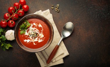 Krema, tohum ve otlarla dolu bir kasede taze domates çorbası servis edilir. Yanında da kaşık ve peçeteyle kahverengi bir zemin üzerinde domates, sarımsak ve maydanoz bulunur.