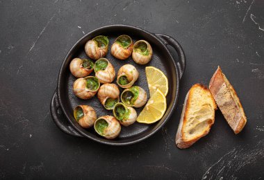 Sarımsak ve maydanozlu tereyağlı Bourgogne salyangozları, ahşap kesme tahtasındaki dökme demir bir tavada servis ediliyor, limon dilimleri ve kızarmış ekmekle birlikte, lezzetli bir Fransız aperatifi.