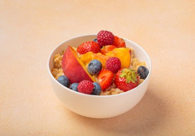 Taze şeftali dilimleri, yabanmersini, çilek ve ahududu ile kaplı beyaz kasede yulaf ezmesi sağlıklı beslenme alışkanlıklarını teşvik eden besleyici ve canlı bir kahvaltı.