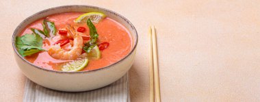 Taze karides, limon kamaları, şifalı otlar ve kırmızı biberlerle süslenmiş ve yanında keten peçetede yemek çubukları bulunan Tayland 'ın popüler bir yemeği olan kremalı Tom yum çorbası bir kâse içinde servis edilir.