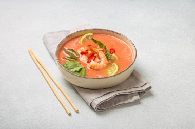 Taze karides, limon kamaları, şifalı otlar ve biberlerle süslenmiş ve yanında keten peçetede yemek çubukları bulunan Tayland 'ın popüler bir yemeği olan kremalı Tom yum çorbası bir kasede servis edilir.