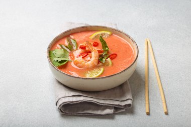 Taze karides, limon kamaları, şifalı otlar ve biberlerle süslenmiş ve yanında keten peçetede yemek çubukları bulunan Tayland 'ın popüler bir yemeği olan kremalı Tom yum çorbası bir kasede servis edilir.