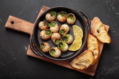 Sarımsak ve maydanozlu tereyağlı Bourgogne salyangozları, ahşap kesme tahtasındaki dökme demir bir tavada servis ediliyor, limon dilimleri ve kızarmış ekmekle birlikte, lezzetli bir Fransız aperatifi.