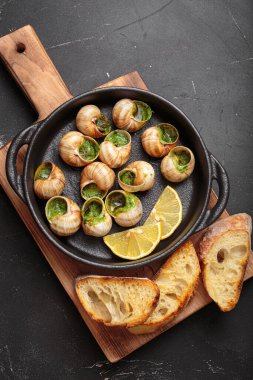 Sarımsak ve maydanozlu tereyağlı Bourgogne salyangozları, ahşap kesme tahtasındaki dökme demir bir tavada servis ediliyor, limon dilimleri ve kızarmış ekmekle birlikte, lezzetli bir Fransız aperatifi.