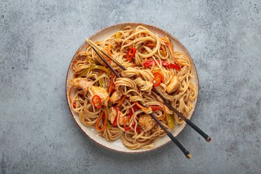 Tavuk eti, havuç, dolma biber ve diğer sebzelerle kızartılmış Udon erişteleri gri arka planda çubuklarla servis edilir ve susam tohumları serpiştirilir.