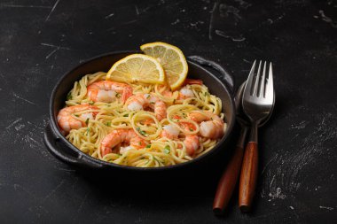 Siyah dökme demir tavada servis edilen karidesli makarna taze limon dilimleri ve siyah kırsal arka planda maydanoz açı görüşüyle süslenmiş.