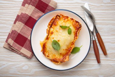 Bol bolonez soslu lazanya, erimiş peynir ve beyaz tabakta taze fesleğen, beyaz tabakta beyaz bir tabakta kareli peçete ve çatalla servis ediliyor.