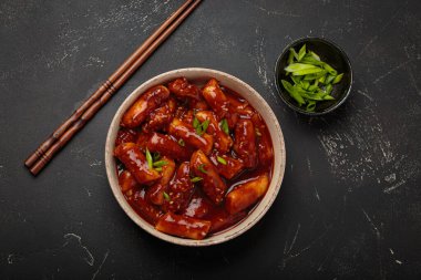 Baharatlı sosla kaynatılmış, yeşil soğan ve susam tohumlarıyla süslenmiş, siyah arka planda servis edilen pirinç kekleri yemek çubukları üst görüşüyle servis edilir. Kore 'nin popüler yemeği.