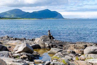 Norway - 07.05.2024: Lone man sitting on rocks and looking at his phone green with mountains, fjord of Norwegian landscape in background clipart