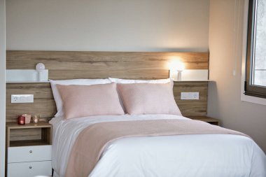 Rahat, modern yatak odası. Yumuşak pembe yastıklar, beyaz yatak örtüsü ve sıcak bir başucu lambası..