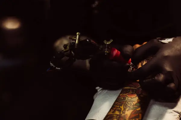 Tattoo artist at work with a machine on skin, close-up, dark moody lighting.