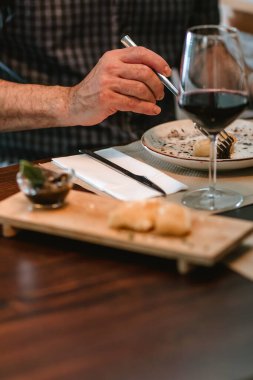 Kareli gömlekli bir adam tabağında bir parça ekmek kesiyor, tahta bir masaya kuruluyor..