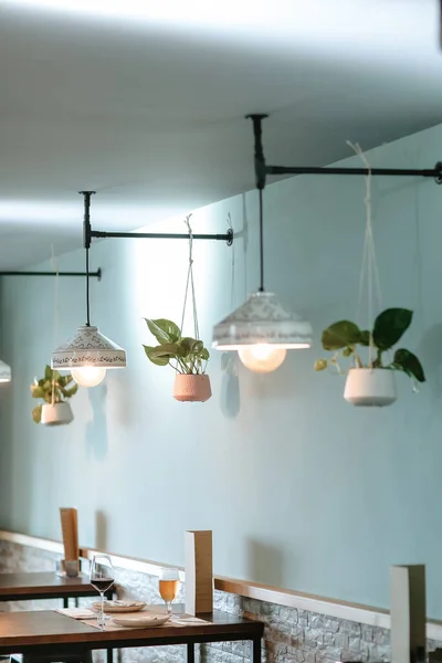 Stylish side view of a bar featuring designer white lamps, hanging plants, dining tables and chairs, with a bar counter in the background.