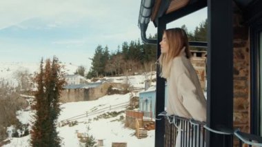 Düşünceli Kış Sabahı: Genç Kadın Modern Balkondan Kar Tozlu Köye ve Tepelere Bakıyor