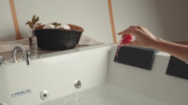 Hand pouring bath salts into a bathtub with faucet and basket of towels nearby. Spa and relaxation setting