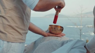 Sound Therapy Session Outdoors with Therapist Using Singing Bowl Over Covered Client in Scenic Ocean Setting