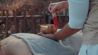 Sound Healing Session Outdoors with Therapist Using Tibetan Singing Bowl Over Client Wrapped in Cozy Blanket