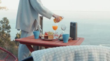 Person Pouring Fresh Orange Juice into Glasses at an Outdoor Breakfast Table with Assorted Food Items Overlooking a Scenic Ocean View