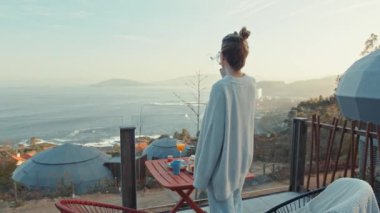 Woman Enjoying Morning Coffee on an Outdoor Terrace with Scenic Ocean View and Breakfast Table Setup
