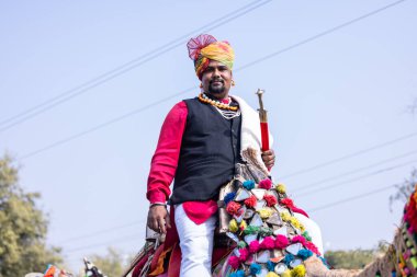 Bikaner, Rajasthan, India - January 2023: Camel Festival, Portrait of an young rajasthani male riding on decorated camel with beard and moustache wearing white traditional rajasthani dress and turban.