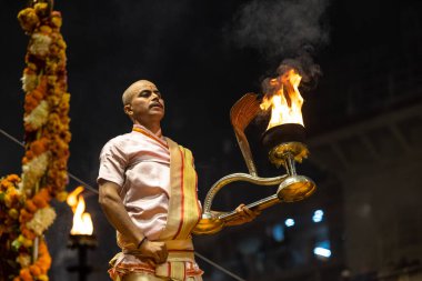 Varanasi, Uttar Pradesh, Hindistan - Kasım 2022: Ganga aarti, genç bir rahibin portresi Daswamedh ghat 'ta akşam üzeri sanatan hindu ayinleriyle geleneksel elbise içinde nehir çetelerini icra ediyor..