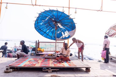 Varanasi, Uttar Pradesh, India - November 2022: Portrait of Unidentified Indian brahmin priest sitting under umbrella on ghat near river ganges in varanasi city. Priests performed rituals on ghats. clipart