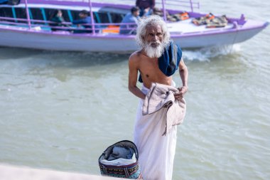 Varanasi, Uttar Pradesh, Hindistan - Kasım 2022: Kimliği belirlenemeyen yaşlı erkek sadhu 'nun portresi gündoğumu sırasında nehir çetelerinin yanında duruyor ve Hindu ayinleri olarak nehir çetelerine kutsal dalış yapıyor..