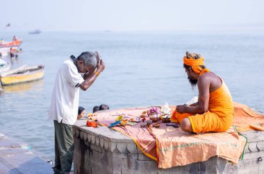 Varanasi, Uttar Pradesh, Hindistan - Kasım 2022: Varanasi şehrinde geleneksel elbise içinde nehir çetelerinin yanındaki Assi ghat 'ta oturan kimliği belirsiz Hintli sadhu baba portresi.