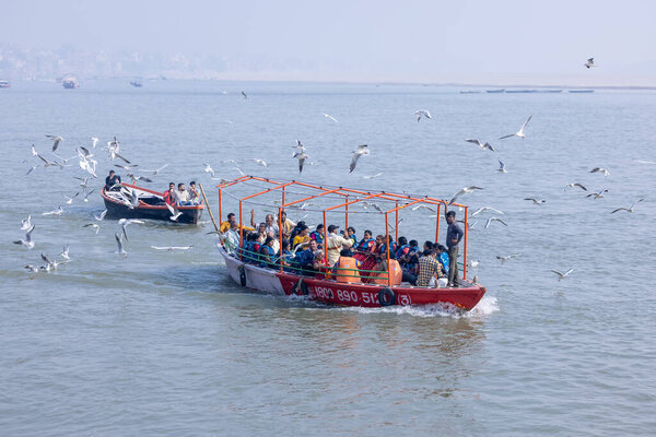 Varanasi, Uttar Pradesh, India - November 2022: Tourists enjoying boat ride in the river ganges along with the herd of sea gulls at varanasi during early morning. Boatman sailing the wooden boat.