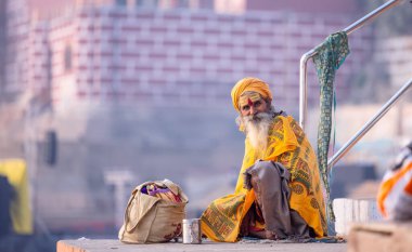 Varanasi, Uttar Pradesh, India - November 2022: Portrait of Unidentified Indian old sadhu baba sitting on colorful stairs on ghats near river ganges in varanasi city in traditional dress. clipart