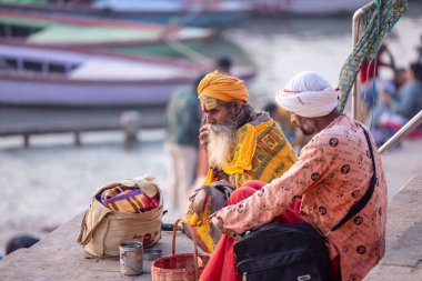 Varanasi, Uttar Pradesh, Hindistan - Kasım 2022: Kimliği belirsiz Hintli yaşlı sadhu Baba 'nın portresi Varanasi şehrinde geleneksel elbise giymiş nehir çetelerinin yanındaki ghat' larda renkli merdivenlerde oturuyor.