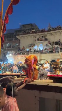 Varanasi, Uttar Pradesh, Hindistan - 20 Mart 2024: Yaşlı bir kadının portresi erkek rahiplerin akşam Ganga aarti performansından önce nehir ganj ghatları üzerindeki diya mumlarını aydınlatıyor.