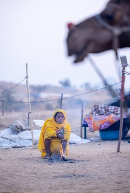 Puşkar, Rajasthan, Hindistan - 24 Kasım 2023: Sarı etnik elbiseli genç bir kadının portresi, kış sabahı şenlik ateşiyle vücudunu ısıtırken itiş deve fuarı sırasında yerde oturuyor.