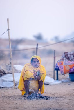 Puşkar, Rajasthan, Hindistan - 24 Kasım 2023: Sarı etnik elbiseli genç bir kadının portresi, kış sabahı şenlik ateşiyle vücudunu ısıtırken itiş deve fuarı sırasında yerde oturuyor.