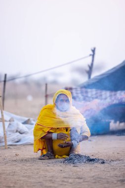 Puşkar, Rajasthan, Hindistan - 24 Kasım 2023: Sarı etnik elbiseli genç bir kadının portresi, kış sabahı şenlik ateşiyle vücudunu ısıtırken itiş deve fuarı sırasında yerde oturuyor.