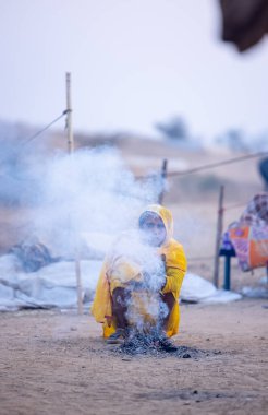 Puşkar, Rajasthan, Hindistan - 24 Kasım 2023: Sarı etnik elbiseli genç bir kadının portresi, kış sabahı şenlik ateşiyle vücudunu ısıtırken itiş deve fuarı sırasında yerde oturuyor.