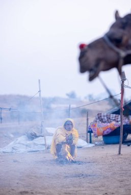 Puşkar, Rajasthan, Hindistan - 24 Kasım 2023: Sarı etnik elbiseli genç bir kadının portresi, kış sabahı şenlik ateşiyle vücudunu ısıtırken itiş deve fuarı sırasında yerde oturuyor.