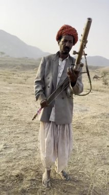 Puşkar, Rajasthan, Hindistan - 24 Kasım 2023: Sarangi müzik enstrümanı çalan geleneksel bir folk müzisyeni erkek sanatçının deve fuarı sırasındaki kum tepeleri üzerindeki videosu.