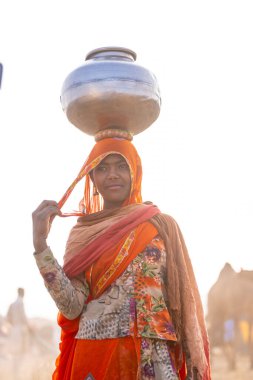 Puşkar, Rajasthan, Hindistan - 24 Kasım 2023: Etnik renkli elbiseli Hintli kadın Pushkar deve panayırında yürürken kafasında çelik su kabı taşıyor. 