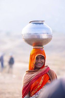 Puşkar, Rajasthan, Hindistan - 24 Kasım 2023: Etnik renkli elbiseli Hintli kadın Pushkar deve panayırında yürürken kafasında çelik su kabı taşıyor. 