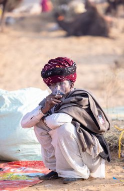 Puşkar, Rajasthan, Hindistan - 23 Kasım 2023: Geleneksel beyaz giysiler ve renkli türbanlı bir adamın portresi Pushkar deve fuarı sırasında çöl panayırında sigara içiyor.