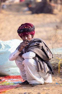 Puşkar, Rajasthan, Hindistan - 23 Kasım 2023: Geleneksel beyaz giysiler ve renkli türbanlı bir adamın portresi Pushkar deve fuarı sırasında çöl panayırında sigara içiyor.