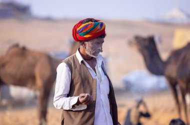 Puşkar, Rajasthan, Hindistan - 23 Kasım 2023: Geleneksel beyaz giysiler ve renkli türbanlı bir adamın portresi Pushkar deve fuarı sırasında çöl panayırında sigara içiyor.