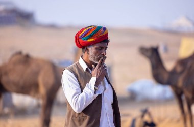 Puşkar, Rajasthan, Hindistan - 23 Kasım 2023: Geleneksel beyaz giysiler ve renkli türbanlı bir adamın portresi Pushkar deve fuarı sırasında çöl panayırında sigara içiyor.
