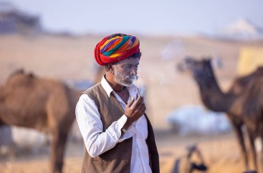 Puşkar, Rajasthan, Hindistan - 23 Kasım 2023: Geleneksel beyaz giysiler ve renkli türbanlı bir adamın portresi Pushkar deve fuarı sırasında çöl panayırında sigara içiyor.