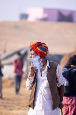 Puşkar, Rajasthan, Hindistan - 23 Kasım 2023: Geleneksel beyaz giysiler ve renkli türbanlı bir adamın portresi Pushkar deve fuarı sırasında çöl panayırında sigara içiyor.